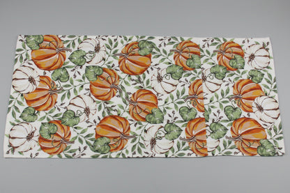 A towel fully opened, featuring a repeating autumn pattern of orange and white pumpkins with green leaves on a white background, laid out on a gray surface.