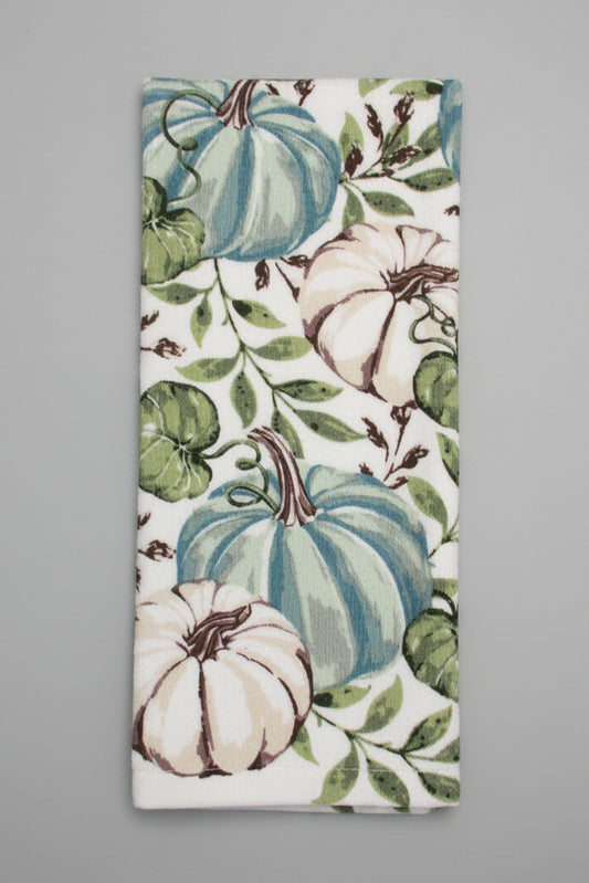 A folded tea towel featuring a vintage-style print of blue-green pumpkins, cream pumpkins, and green leaves on a white background, laid flat on a dark gray surface.