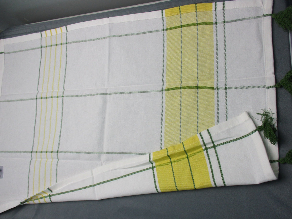 Back side of white cotton kitchen towel with green and yellow striped plaid patterns, featuring green tassels on one end, laid out on a dark gray surface.