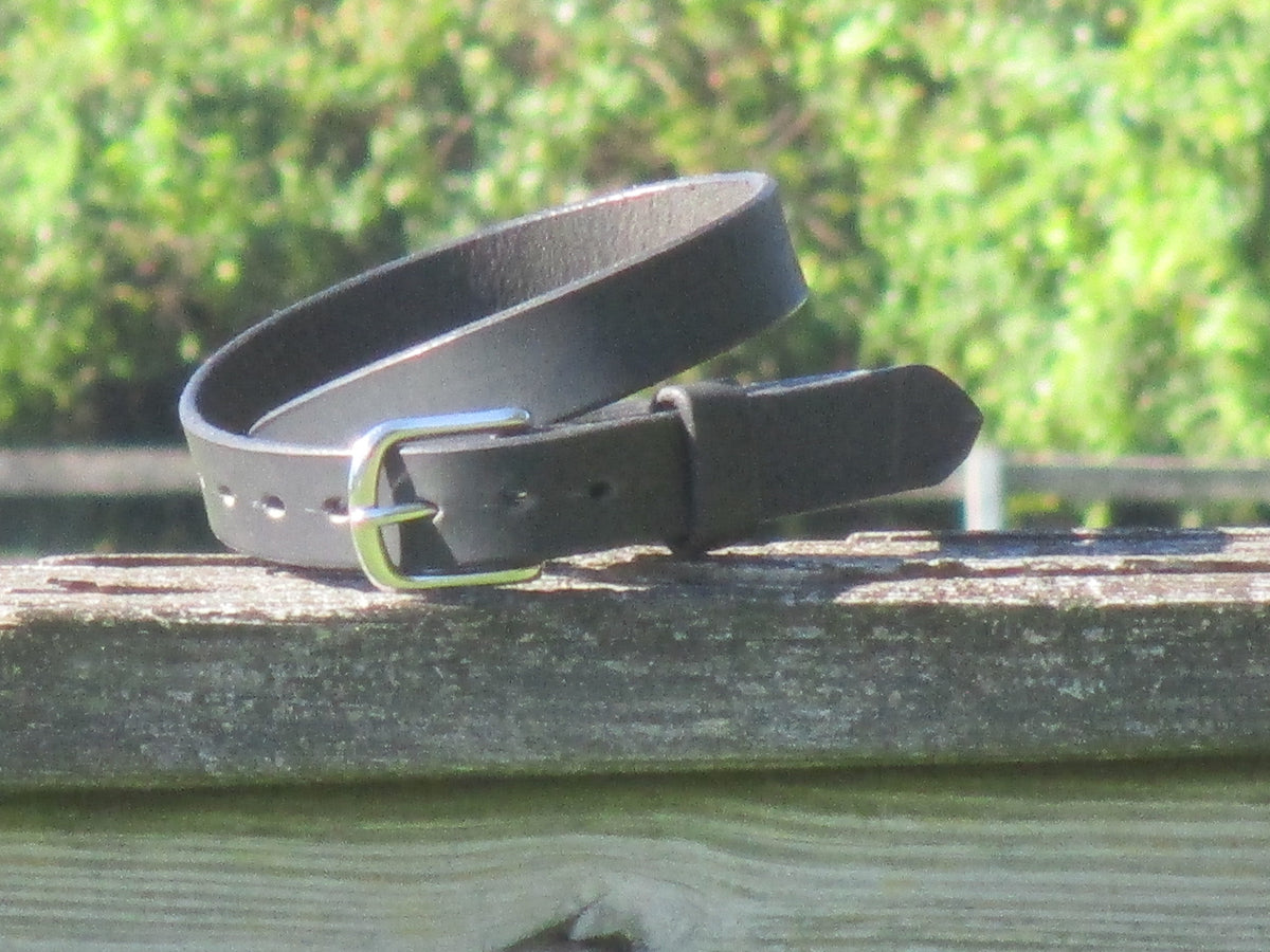 Water Buffalo Black leather belt with a Stainless Steel buckle on a wooden surface with a blurred green background